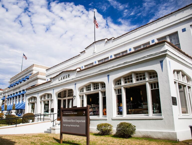 The white Lamar Bathhouse on Bathhouse Row in Hot Springs, Arkansas, with a National Park Service sign in front and the Buckstaff Bathhouse behind it